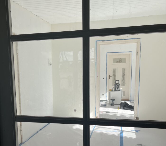 Ett nyrenoverat rum med vita väggar och tak, sett genom en glasdörr. Golvet är täckt med skyddspapp. Verktyg och färgburkar syns.