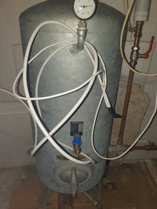 Hydrofor i källare med tryckmätare och anslutna rör, inklusive vita plastslangar och ventil.