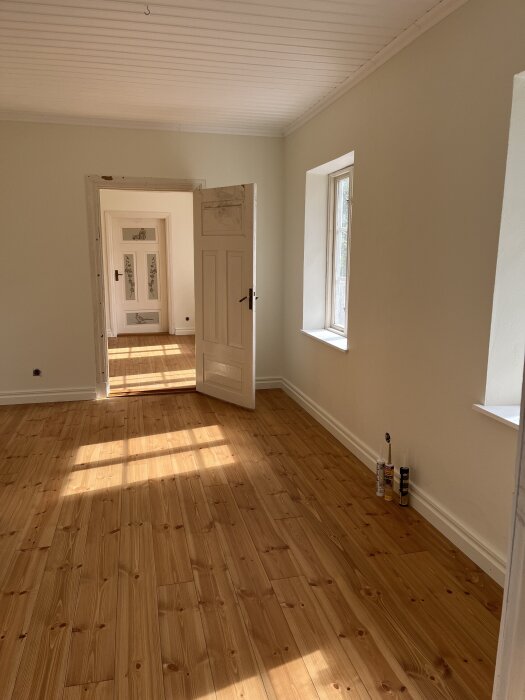 Ett nyrenoverat rum med vitmålade väggar och tak, samt ett öppet fönster och träparkettgolv badande i solljus.