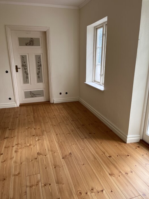 Ljusa väggar och trägolv i ett rum med en dörr dekorerad med illustrationer, fönster med fönsterbräda och golvlister.