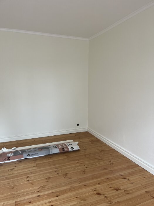 Nymålat rum med vita väggar och trägolv, visar nyansskillnad i hörnet. Förpackningar ligger på golvet. Målningen är nästan klar för inflytt.