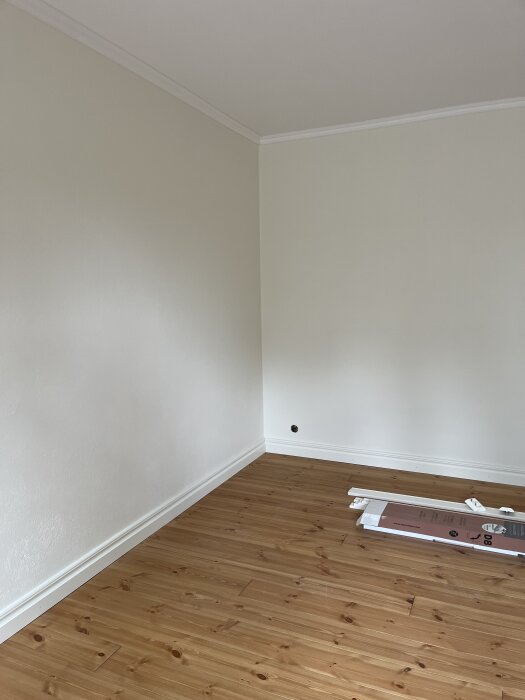 Ett ljust rum med vita väggar och ett träfärgat golv. En kartong och några föremål ligger på golvet. En nyansskillnad syns i hörnet.