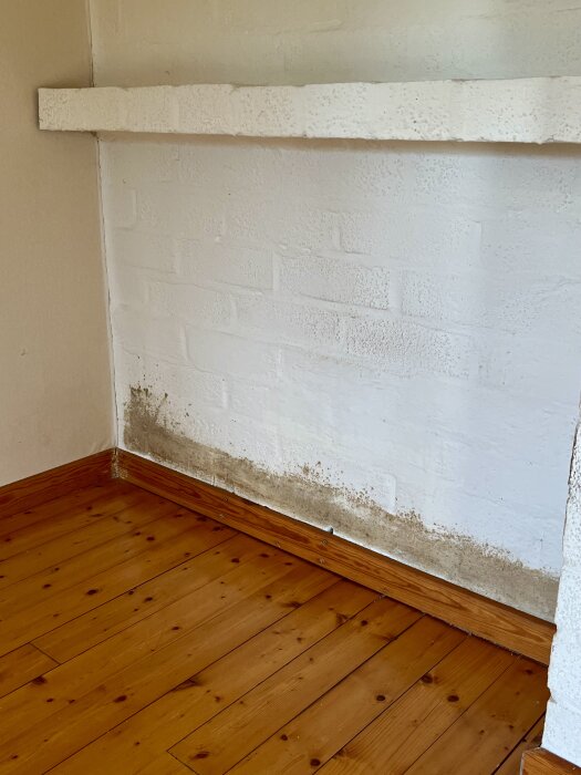 Vitmålad tegelvägg vid hörna med golvlister i trä. Mörka fläckar syns längs väggen nära golvet.