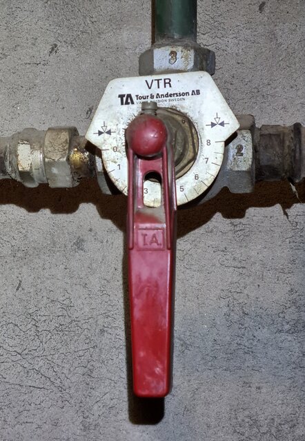 Shuntventil med röda vredet och markerade anslutningar, modell från Tour & Andersson AB, kopplad till rör på betongvägg.
