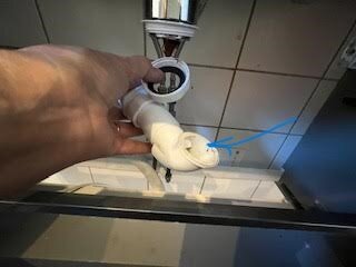 En hand håller i ett vitt VVS-rör under ett handfat. Rörets krök markeras med en blå pil.