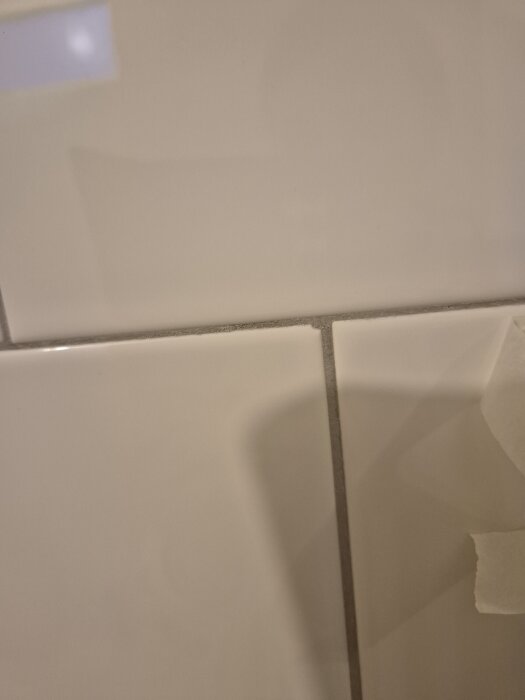 Vägg med kakelplattor där hörn saknas på vissa och en platta är intryckt, med ojämn list runt ett fönster.