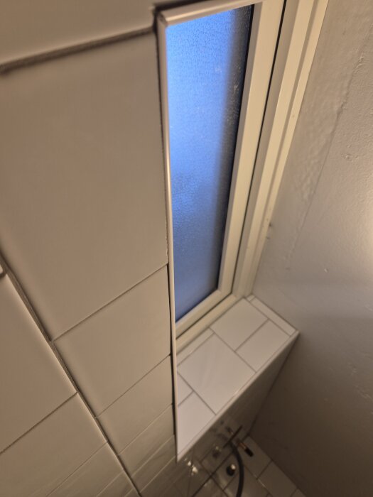 Vägg med vita kakelplattor vid fönster, en platta skev och hörn saknas på vissa plattor, ojämn list runt fönster.