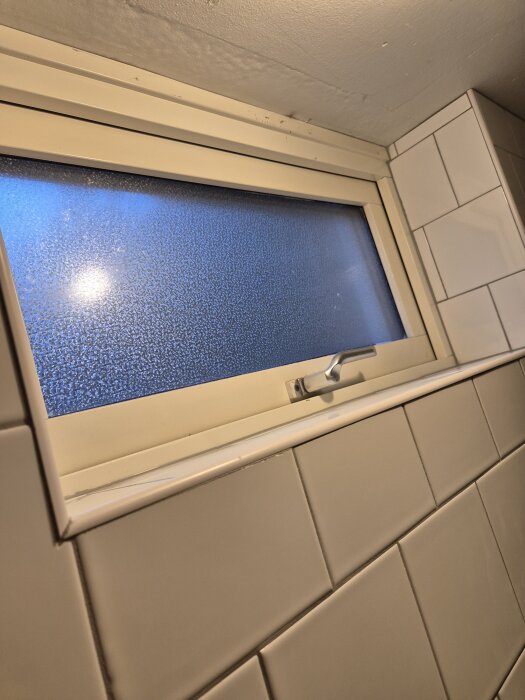 Kakelvägg runt frostat fönster med ojämna plattor och hörn saknas på vissa plattor. Ojämn list runt fönstret.