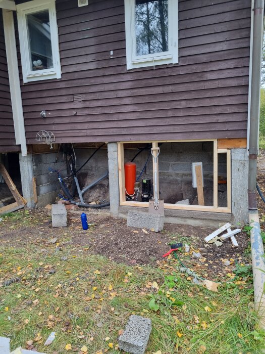 Renoverat pumphus med öppning i brun träfasad, lecablock och verktyg på marken; diskuteras val av panelmaterial.