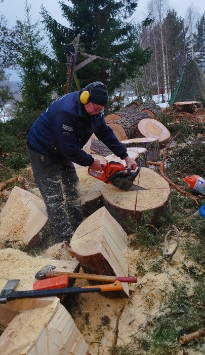 Person kapar ved med motorsåg bland trädstammar och sågspån, utrustad med hörselskydd och arbetskläder i en skogsmiljö.