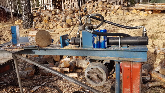 Vedklyv med stock och två blå burkar på hyvelbänk. Stubbar och vedträn i bakgrunden, utomhusmiljö med björkar.