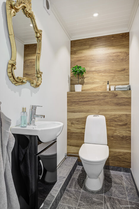 Klassisk liten toalett med vit porslinstoalett, träpanel på väggen, svart handfatsskåp, guldspegel och grått klinkergolv.