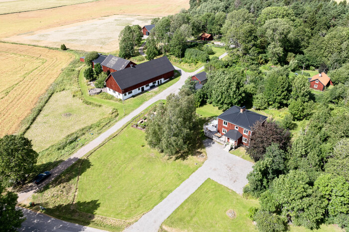 Flygfoto över en gård med röda hus omgivna av träd och åkrar, en grusväg leder till huvudbyggnaden och odlingsträdgården.