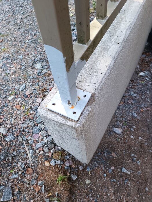 Smidesräcke monterat på L-stöd vid källarnedgång, fäste med platta och bultar syns, omgiven av grus och jord.