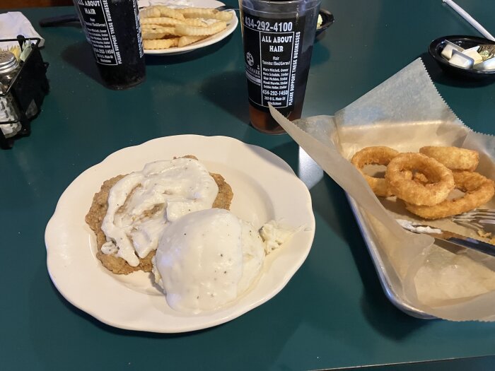 Tallrik med friterad kyckling och potatismos täckt med sås, bredvid en tallrik med lökringar och krusiga pommes frites.