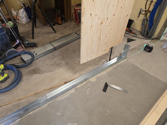 Byggarbetsplats med en stålregel, träskiva, laserverktyg och mätverktyg på golvet. Verktyg och material syns runtom.