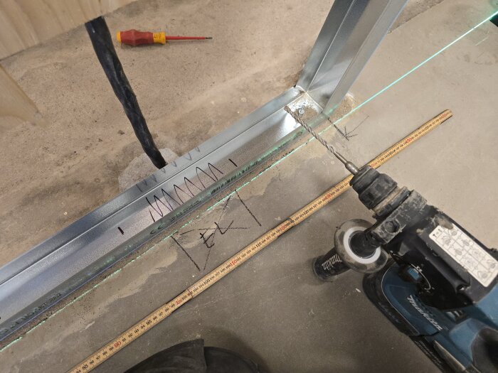 Borrmaskin borrar hål i golvet bredvid stålskena, tumstock och verktyg, förbereder montering av takskena.