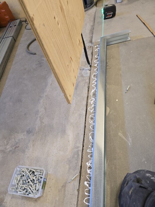 Stålregel monteras med fogmassa intill träregel, lasernivå i bakgrunden. Skruvar och plugg syns på golvet, samt sladdar och verktyg.