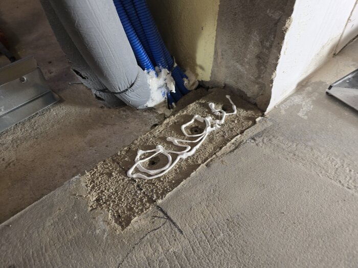 Rördragning med el- och ventilationsrör i en byggarbetsplats hörn, synlig fogmassa applicerad på betonggolvet.