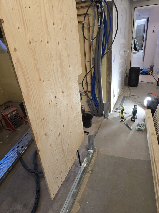 Byggplats med träpanel, verktyg och stålskena. Synliga rör och kablar på vägg; pågående renoveringsprojekt med fokus på montering och eldragning.