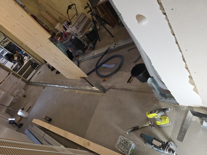 Byggarbetsplats med delvis byggd vägg, verktyg som borrmaskiner och skruvar på golvet, trä- och stålskenor runtomkring.