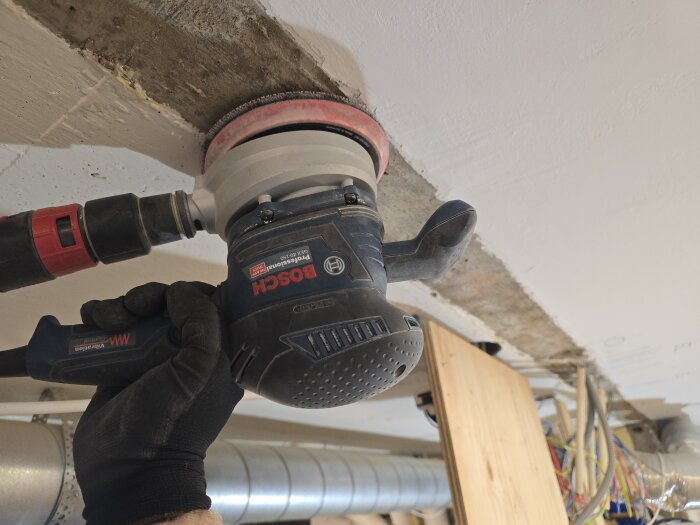 Person slipar blåbetongvägg med en Bosch slipmaskin, iförd handske, under renovering. Ledningar och ventilationsrör syns i bakgrunden.