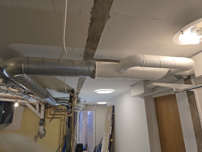 Ventilationsrör i taket i ett rum under renovering, med diverse synliga rör och ledningar i bakgrunden samt en dörr till höger.