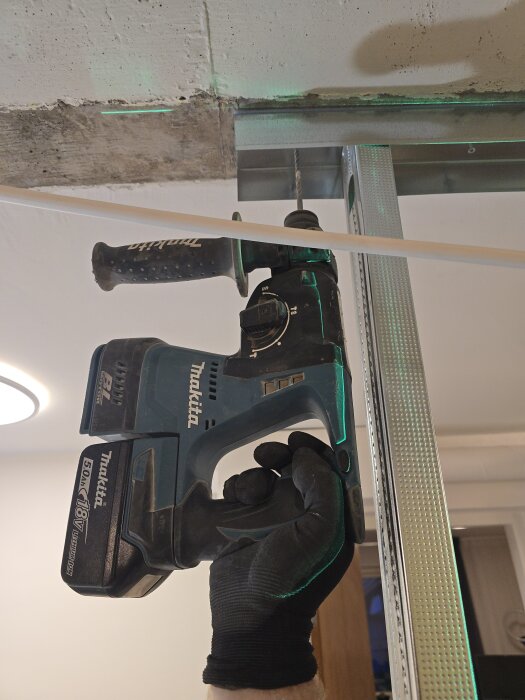 En person borrar i taket med en blå borrmaskin, stålregel och grön laserlinje synliga. Glödlampsarmatur lyser i bakgrunden.