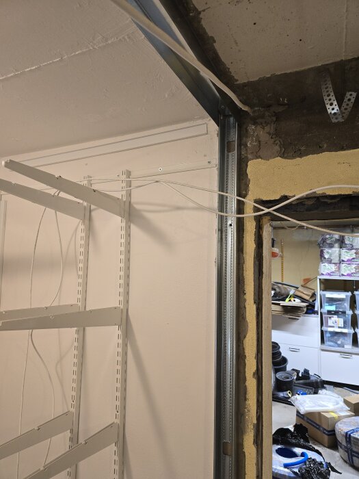 Vägghörna med stålskena och kabeldragning i ett garage under konstruktion. Hyllsystem ses på väggen. Verktyg och material på golvet.