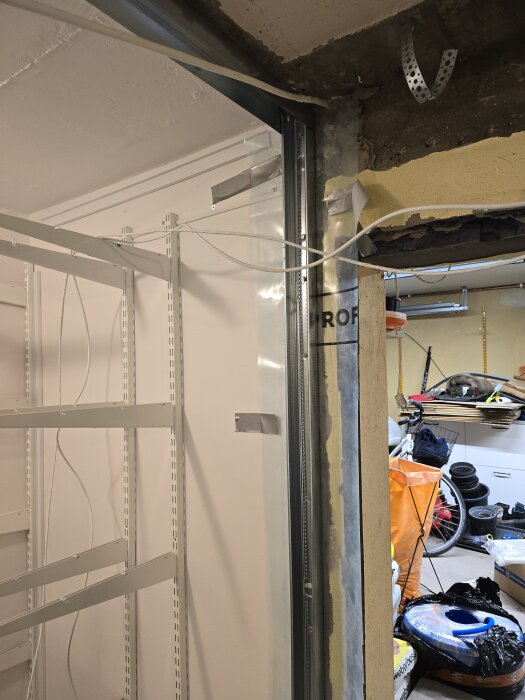 Montering av stålskena med elledningar och plast i en ofärdig väggsektion, omgiven av byggmaterial och verktyg i ett garage.