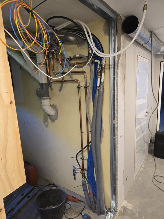 Vägg under konstruktion med exponerade rör och elledningar. Stålskenor och plast användes kring dörrparti. Byggmaterial och verktyg syns på golvet.