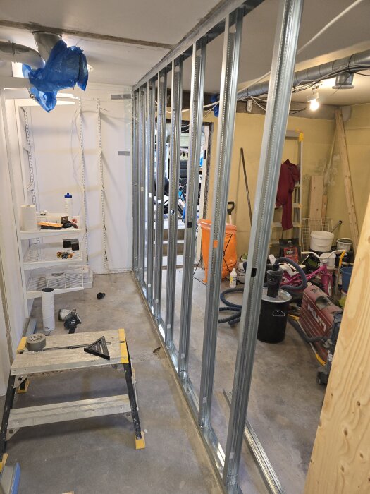 Stålskenor monterade för innerväggsbygge i förrådsutrymme, med verktyg och material spridda på golvet samt arbetsbänk i förgrunden.