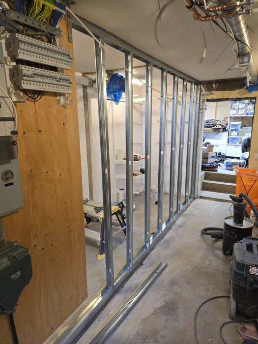 Montering av stålreglar längs en vägg med synliga elinstallationer och verktyg i en pågående renovering av förråd, med byggmaterial på golvet.