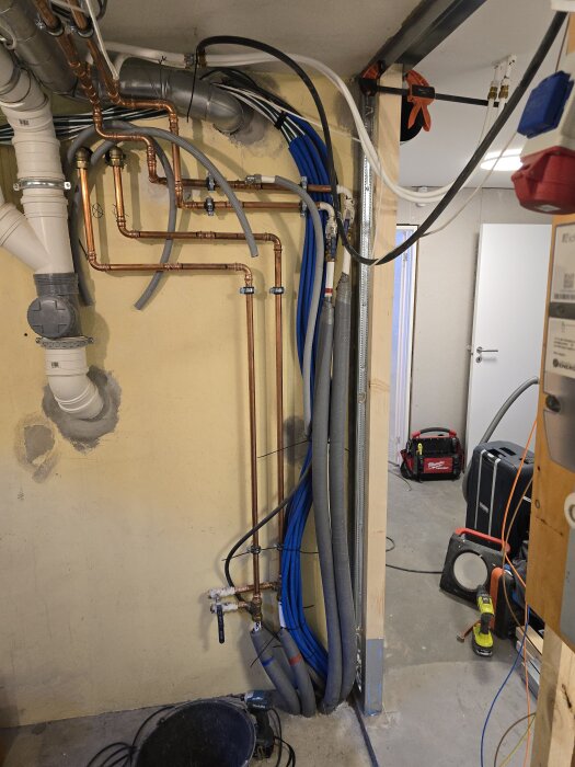 Vägg med kopparrör, elkablar och avloppsrör i en byggzon; verktyg och utrustning synliga på golvet.