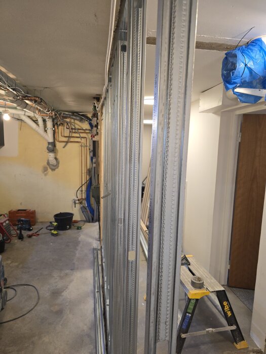 Byggprojekt med stålprofiler och reglar mot en vägg, omgivet av verktyg och ledningar i ett rum under renovering.