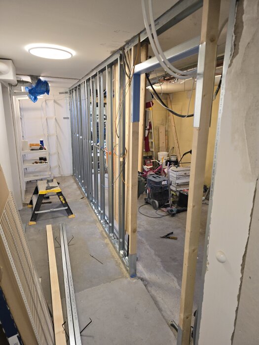 Byggarbetsplats med uppsatta stålreglar i ett rum under konstruktion, verktyg och material synliga på golvet, inklusive en arbetsbänk och kablar.