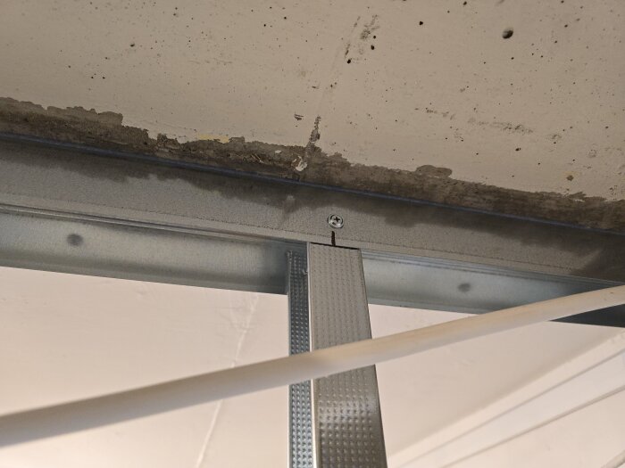 Stålskena monterad i taket med en vertikal regel inskjuten i skenan. Takskenan är fäst med en skruv mot ett betongtak.