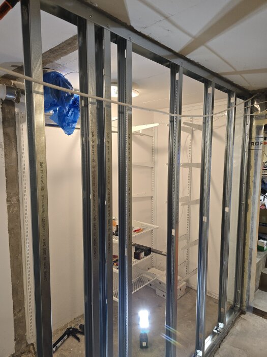 Stålreglar installerade på väggen i ett rum, med verktyg på golvet; plastskydd synligt till vänster, lysrör upplyser arbetsområdet.