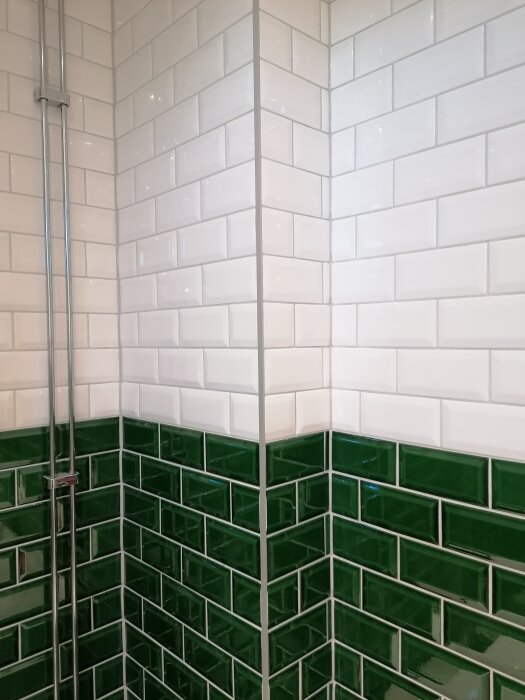 Hörn med vit och grön kakelvägg, borstad aluminiumlist i mitten. Silverfärgade vattenrör syns på vänster sida.