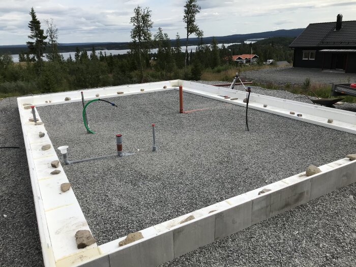 Byggplats för fritidshus med synliga avlopps- och tilluftsrör. Orange rör sticker upp, omgiven av grus och isoleringsmaterial, med träd och sjö i bakgrunden.