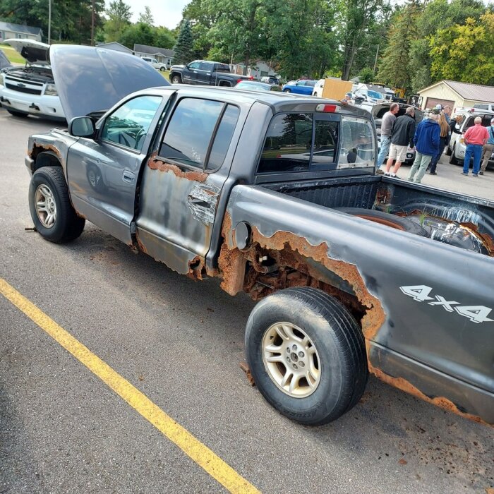 En rostig pickup med 4x4-märkning och synlig skada på karossen, omgiven av människor och andra fordon på en parkering.