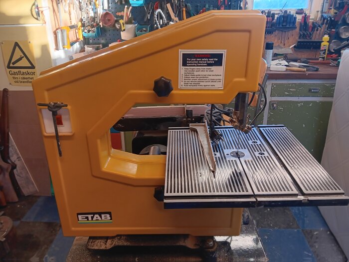 Gul bandsåg av märket Etab i en verkstad, medågblad och en varningsskylt synliga. Verktyg och skyltar i bakgrunden.