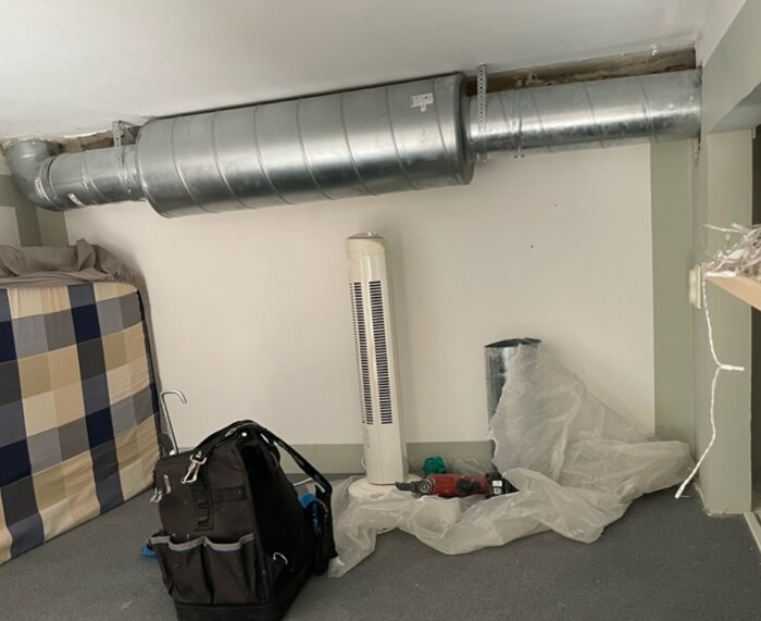 Exponerat ventilationsrör i taket på ett loft med verktyg och material nedanför, inklusive en verktygsväska och ett plastskynke.