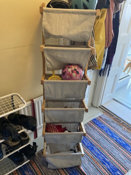 Stående hylla med fyra tygkorgar, fylld med föremål, stående bredvid en skoställning i en hall med randig matta.