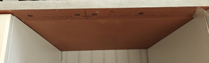 Skyddsplast på bänkens undersida har börjat lossna vid kanten; brun skyddsplast med tryckta markeringar syns ovanför vita skåpväggar.