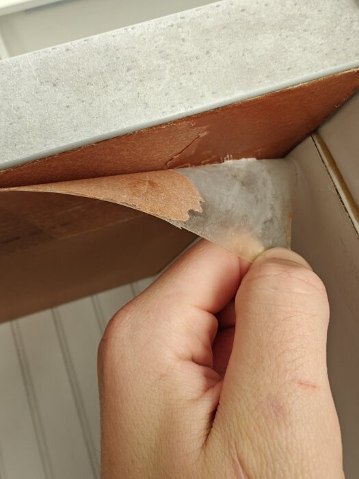 Hand som drar i skyddsplast på en bänkskiva där översta lagret har börjat lossna.