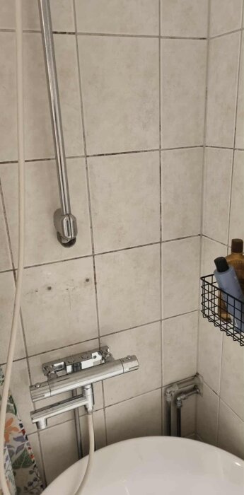 Duschhörna med kakelväggar, duschmunstycke och synliga rör vid duschplatsen. Golvnära rör och produkter på vägghylla.