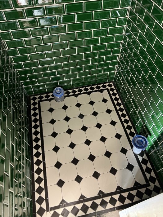 Kaklat badrum med gröna väggar och svartvita plattor på golvet. Två burkar placerade på golvet.
