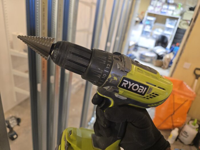 En Ryobi borrmaskin med stegborr hålls i ett bygge omgiven av stålreglar, verktyg och material.