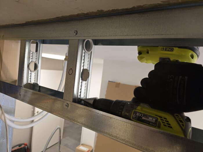 Borrning i stålregel med en sladdlös Ryobi-borrmaskin visas sett uppåt mot ett tak; flexrör och plastringar syns vid borrade hål.